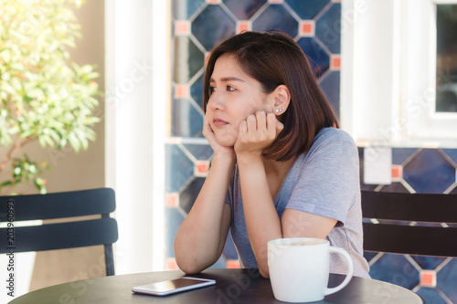 Cheerful asian young woman sitting in cafe drinking coffee and using smartphone for talking, reading and texting. Attractive asian woman holding a cup of coffee while looking on her phone.