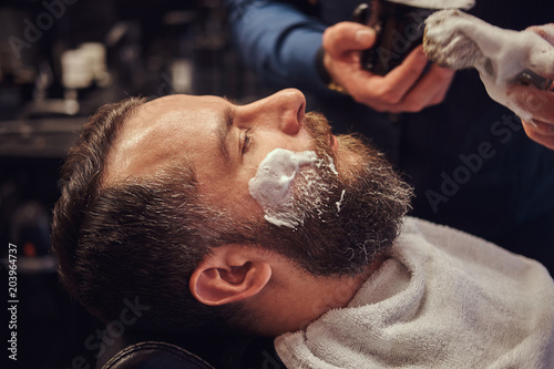 Master hairdresser prepares the face for shaving and smears the face with foam in a hairdressing salon.