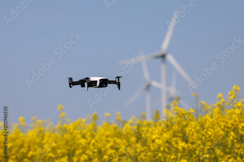 Mini drone floating above rapeseed field surveilling the wind turbines, recording videos and taking pictures. New and modern guardian of big companies. Blurred background