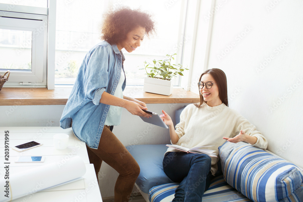 Two girls are studying together. The girl in blue shirt is showing the tablet to the girl is white sweater. The last one is holding a notebook. Girls are laughing. Two girls are studying together. The girl in blue shirt is showing the tablet to the girl is white sweater. The last one is holding a notebook. Girls are laughing.