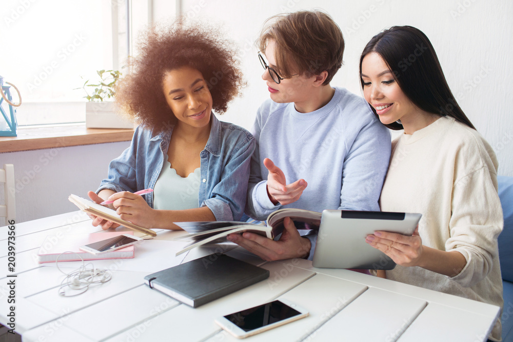 Three smart students are sitting together and studying. Guy wears glasses and looking to the girl's phone while the girls in are looking to the journal that guy has. Three smart students are sitting together and studying. Guy wears glasses and looking to the girl's phone while the girls in are looking to the journal that guy has.