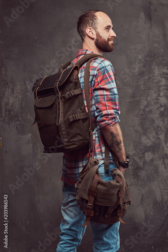 Portrait of a handsome tattooed traveler in a flannel shirt with a backpack, standing in a studio.