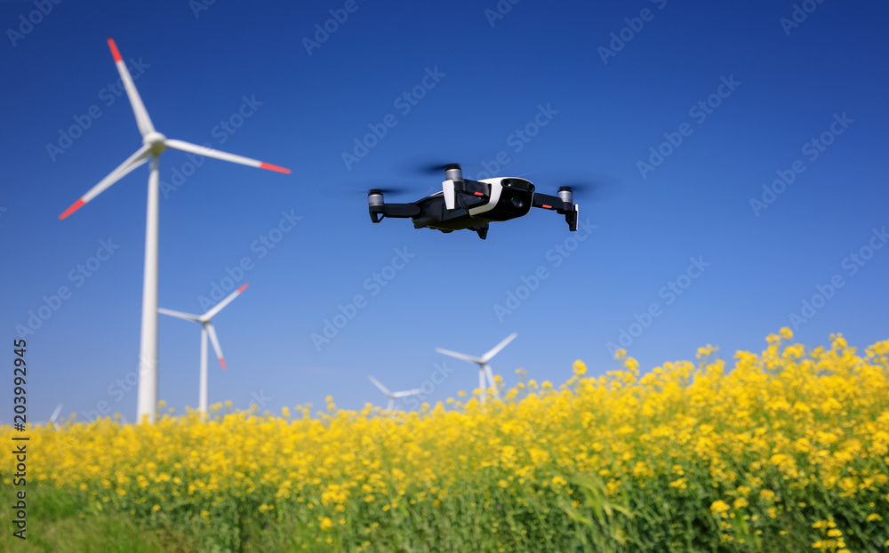 Inspection drone with eolian turbines behind. Rapeseed field in bloom. Renewable energy. - obrazy, fototapety, plakaty 