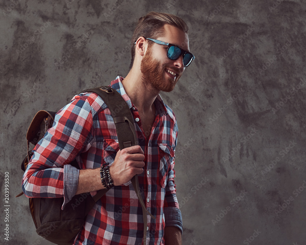 Handsome stylish redhead traveler in a flannel shirt and sunglasses with a backpack, posing in a studio on a gray background.