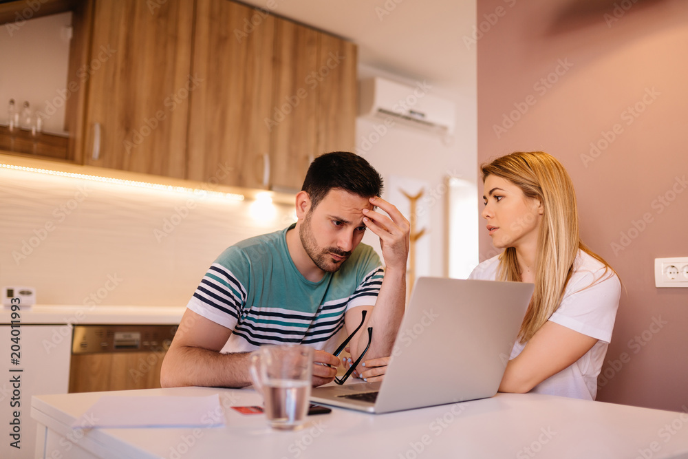 Worried couple checking bank account balance online while sitting in a modern kitchen
