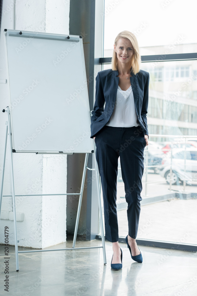 stylish blonde businesswoman standing with hands in pockets and smiling at camera in office