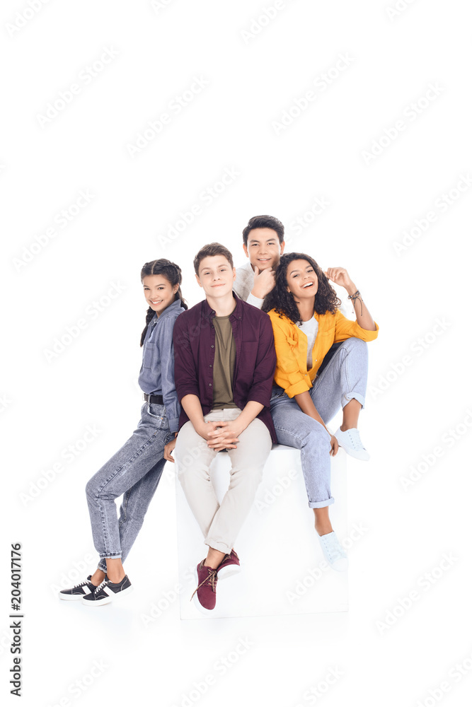 group of happy multiethnic students sitting on white cube isolated on white
