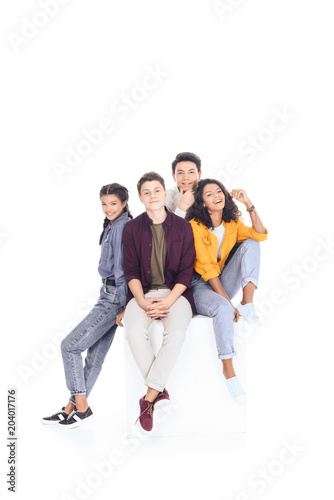 group of happy multiethnic students sitting on white cube isolated on white