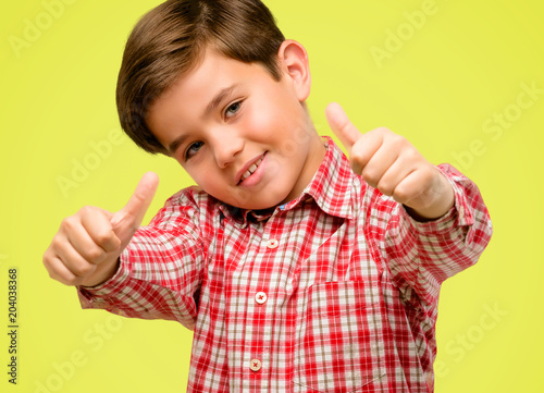 Handsome toddler child with green eyes stand happy and positive with thumbs up approving with a big smile expressing okay gesture over yellow background