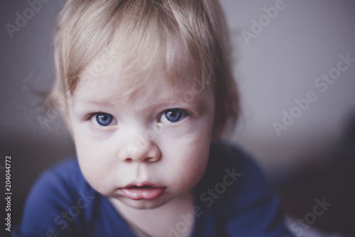 Cute little blond kid with blue eyes in blue clothes looking right into the camera. Close up. Imitation of film grain.