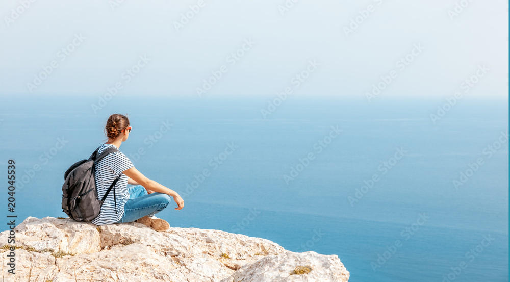 A girl traveler sits on a rock and admires the blue boundless sea, freedom, travel, unity with nature