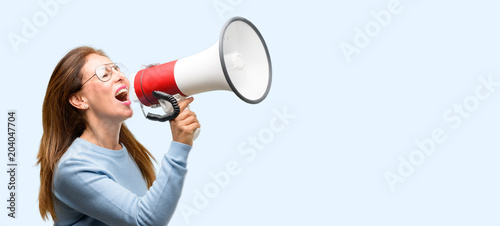 Middle age woman wearing wool sweater and cool glasses communicates shouting loud holding a megaphone, expressing success and positive concept, idea for marketing or sales isolated blue background