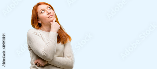 Beautiful young redhead woman thinking and looking up expressing doubt and wonder isolated over blue background