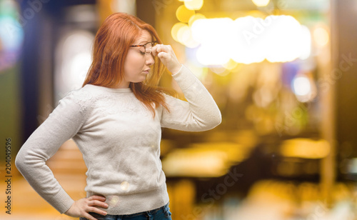 Beautiful young redhead woman with sleepy expression, being overworked and tired, rubbes nose because of weariness at night © Krakenimages.com