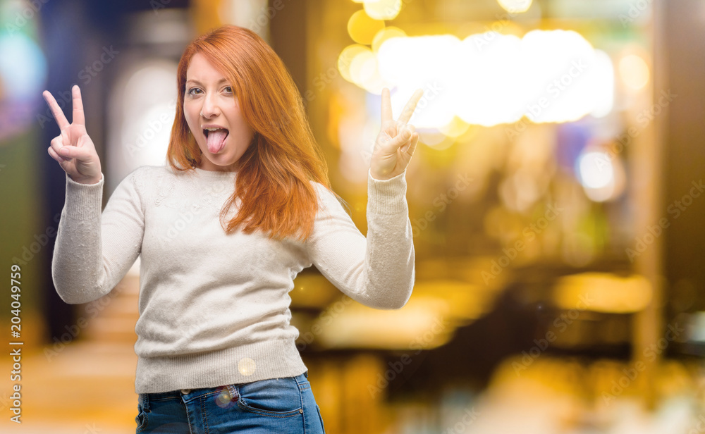 Beautiful young redhead woman looking at camera showing tong and making victory sign with fingers at night