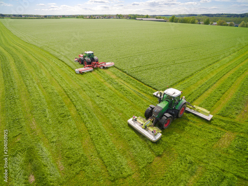   Aerial view of a farmer in a modern tractor mowing a green fresh grass field on a sunny day with blue sky.   