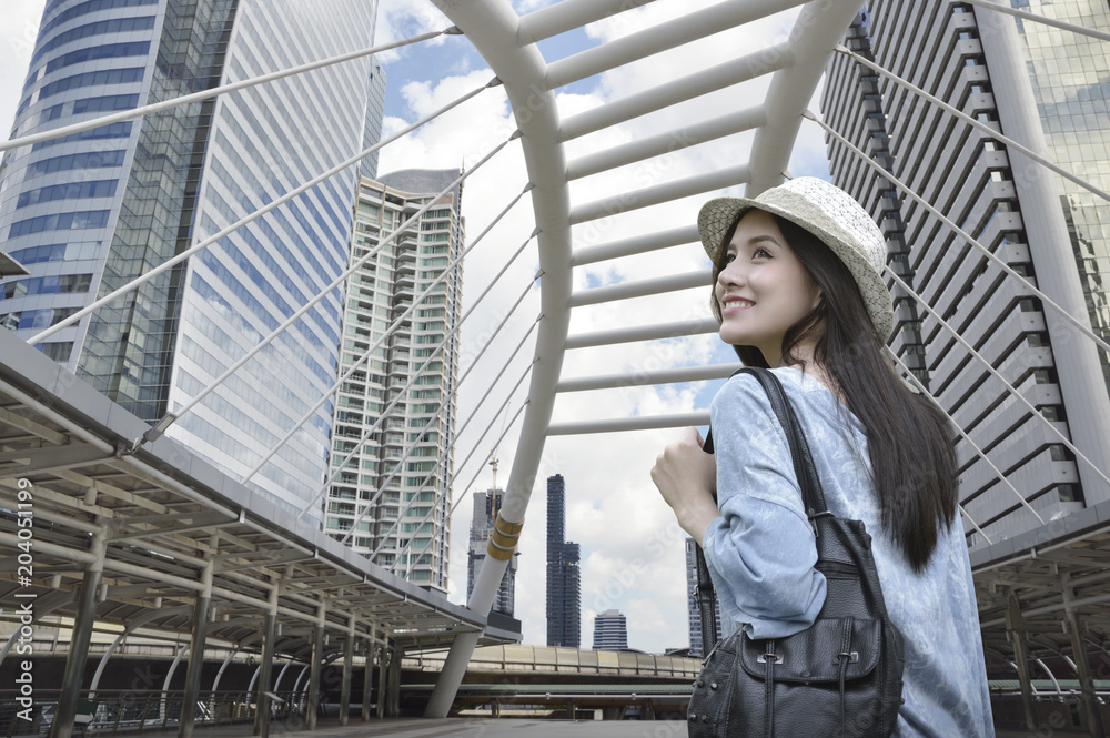 Young asian woman traveler with a backpack on her shoulder walking on pathway bridge over modern city tower in Bangkok, Thailand, Travel holiday relaxation concept