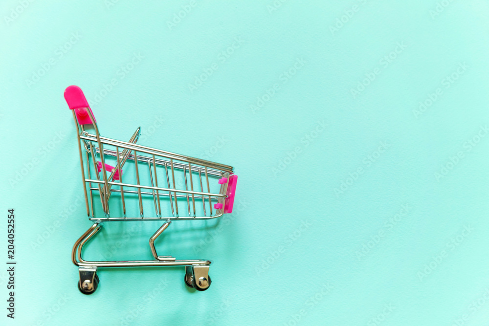 Small supermarket grocery push cart for shopping toy with wheels and pink plastic elements on blue pastel color paper geometric flat lay background. Concept of shopping. Copy space for advertisement Small supermarket grocery push cart for shopping toy with wheels and pink plastic elements on blue pastel color paper geometric flat lay background. Concept of shopping. Copy space for advertisement