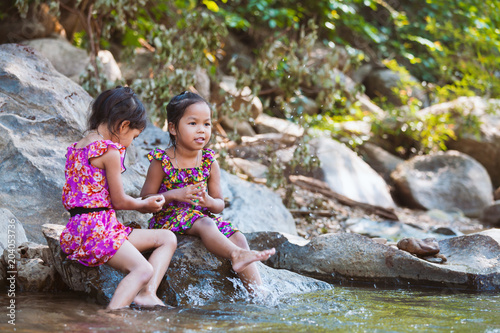 Two asian little child girls sitting on the rock and playing water in the riverside together in summer time with fun and happiness