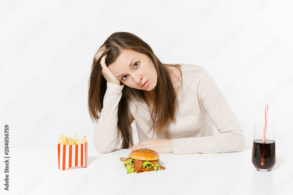 Sad tired woman put hand on head sitting at table with burger, french fries, cola in glass bottle isolated on white background. Proper nutrition or American classic fast food. Area with copy space. Sad tired woman put hand on head sitting at table with burger, french fries, cola in glass bottle isolated on white background. Proper nutrition or American classic fast food. Area with copy space.