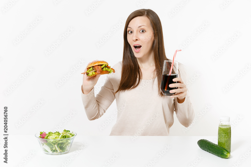 Woman at table with green detox smoothies, salad in glass bowl, cucumber, burger, cola in bottle isolated on white background. Proper nutrition, healthy lifestyle, classic fast food, dieting concept.