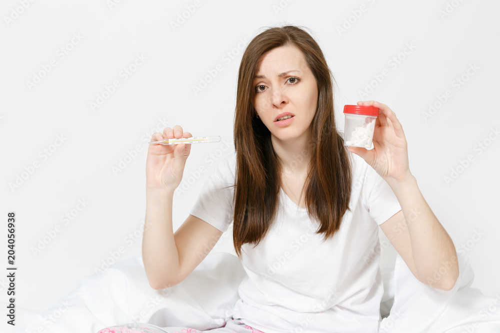 Young sick tired sad woman in pajamas sitting on bed, holding clinical thermometer with high fever temperature, bottle with white pills isolated on white background. Female feeling bad, time at home. Young sick tired sad woman in pajamas sitting on bed, holding clinical thermometer with high fever temperature, bottle with white pills isolated on white background. Female feeling bad, time at home.