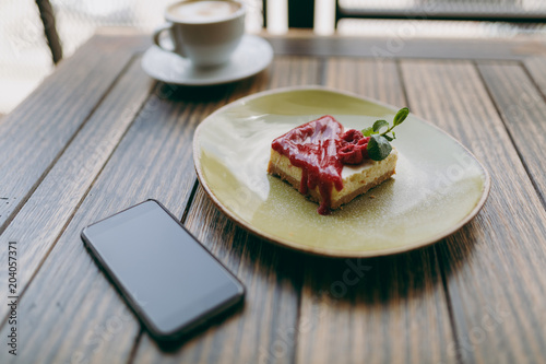 Close up of cake with raspberry sauce on plate, cup of cappuccino, mobile phone with blank empty screen lying on wooden table in coffee shop, cafe, restaurant. Overhead top view. Lifestyle concept.