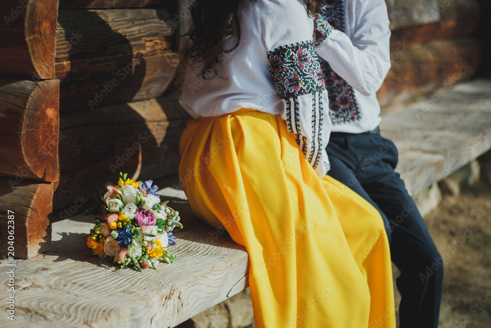 Wedding couple is sitting on the wooden bench on sunny day. Bride in yellow skirt and groom in embroidered shirt. Traditional ukrainian engagement ceremony. Bright bouquet is lying nearby.
