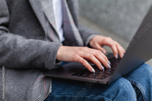 Young stylish fashion blogger using laptop for work while sitting outside on a bench, crop of beautiful woman hands typing on portable computer for distance job, businesswoman working on pc in park