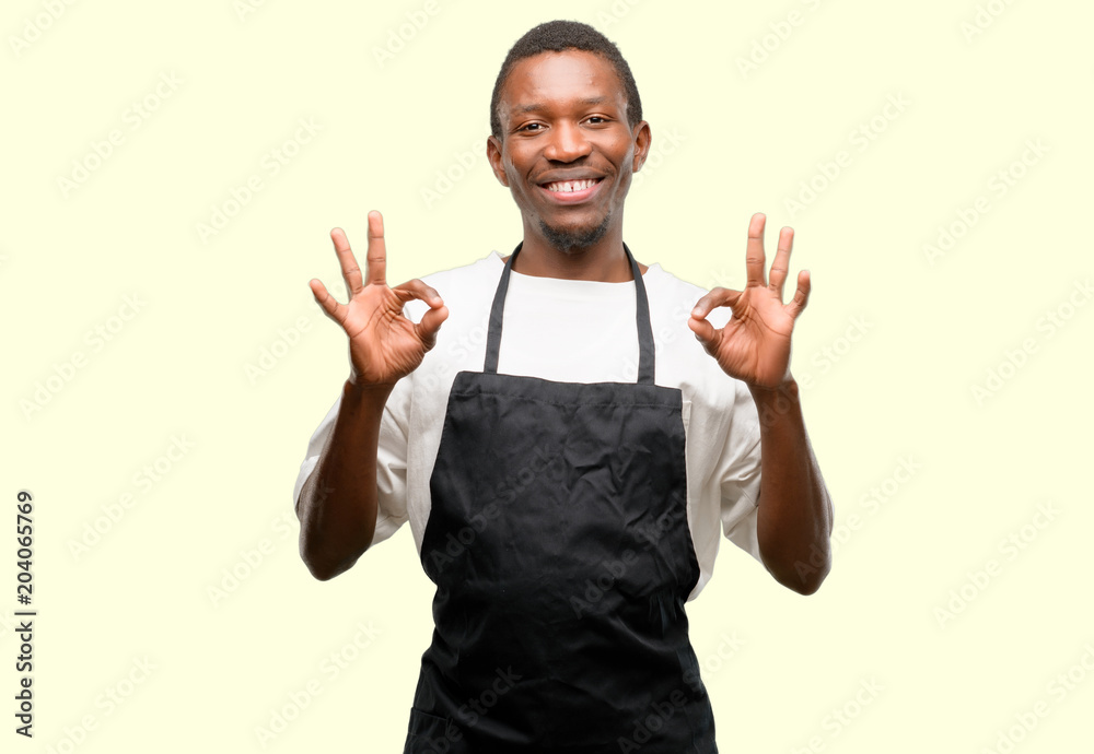 African man shop owner wearing apron doing ok sign gesture with both hands expressing meditation and relaxation African man shop owner wearing apron doing ok sign gesture with both hands expressing meditation and relaxation