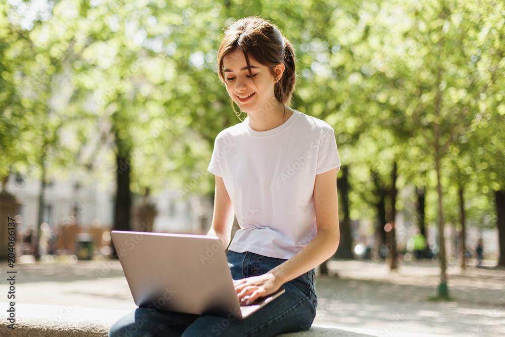 Young blogger using wireless connection to watch video in the park and talk via Skype with friends, beautiful student typing on portable computer for distance job and study while sitting outside Young blogger using wireless connection to watch video in the park and talk via Skype with friends, beautiful student typing on portable computer for distance job and study while sitting outside