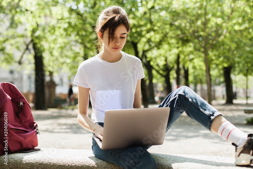 Hipster smiling girl dressed in white t-shirt and jeans studying at laptop in the park near her university, pretty female student preparing for exam via her pc, freelanser working in the fresh air photo