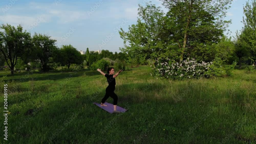 Aerial drone view of a young woman practising yoga outdoors on the grass in the park on summer evening during sunset. Drone flying down Aerial drone view of a young woman practising yoga outdoors on the grass in the park on summer evening during sunset. Drone flying down