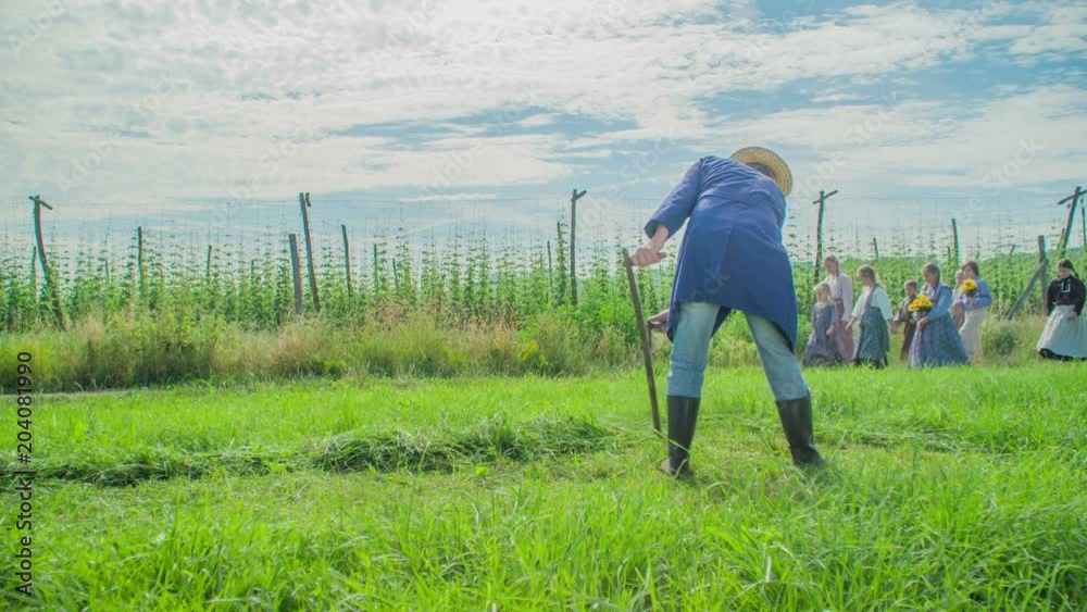 A farmer is cutting grass with a scythe and then women and girls wearing traditional clothes are passin by. A man lifts his hat up when they approach him to show them respect. A farmer is cutting grass with a scythe and then women and girls wearing traditional clothes are passin by. A man lifts his hat up when they approach him to show them respect.