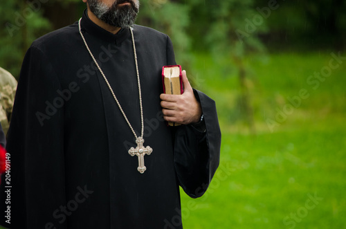 An  unrecognizable orthodox priest with beard in black cloth with silver cross holding a Holy Bible 