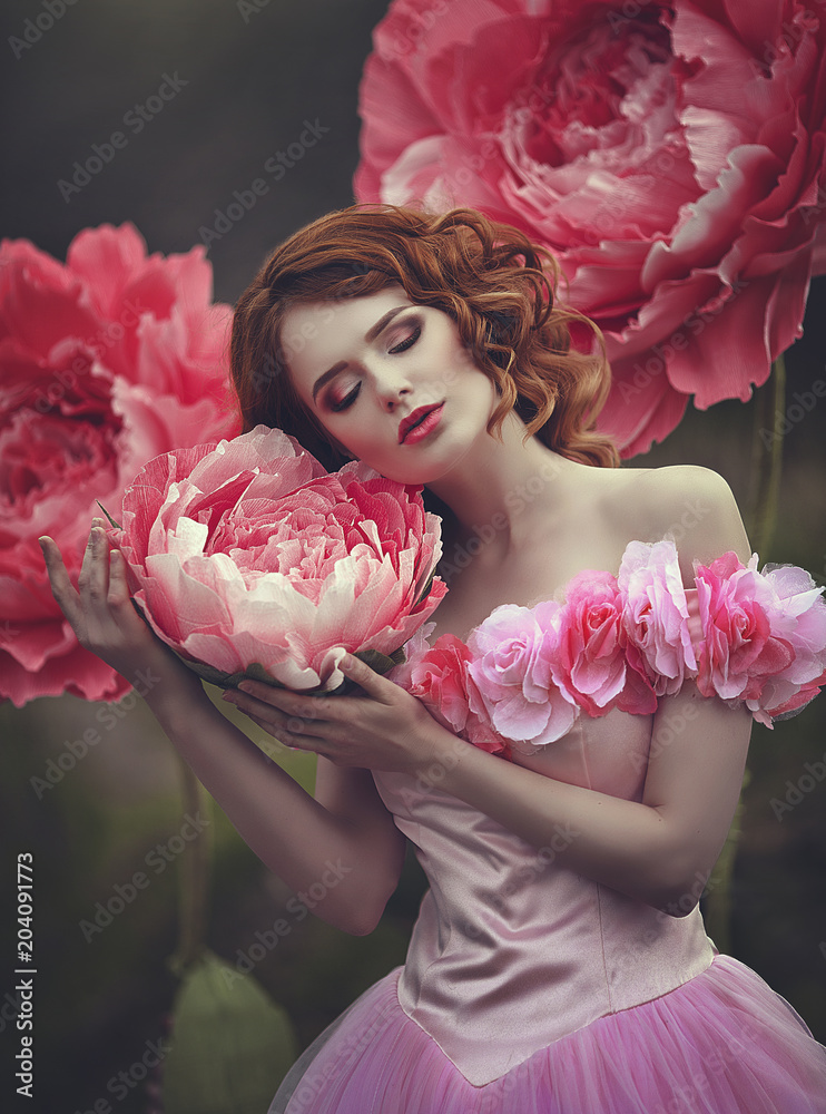 Beautiful sensual girl with red hair in a fairy pink dress posing near  giant peony pink flowers. The girl is a flower princess. Stock Photo