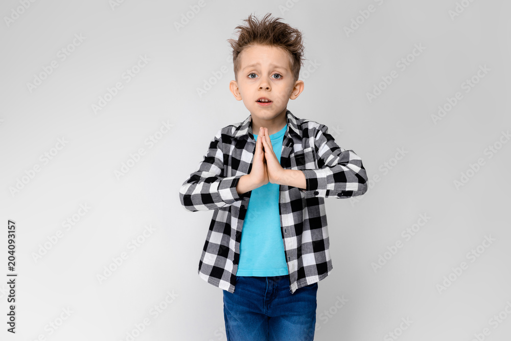 A handsome boy in a plaid shirt, blue shirt and jeans stands on a gray background. The boy folded his arms over his chest. The boy folded his palms against his chest A handsome boy in a plaid shirt, blue shirt and jeans stands on a gray background. The boy folded his arms over his chest. The boy folded his palms against his chest