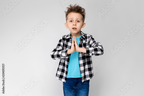 A handsome boy in a plaid shirt, blue shirt and jeans stands on a gray background. The boy folded his arms over his chest. The boy folded his palms against his chest