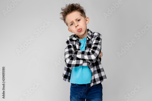 A handsome boy in a plaid shirt, blue shirt and jeans stands on a gray background. The boy folded his arms over his chest. The boy shows the language