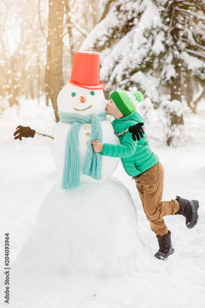 Happy little boy kissing snowman at sunset Happy little boy kissing snowman at sunset