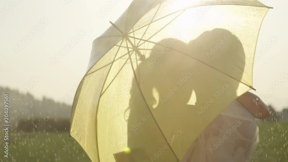 SLOW MOTION, CLOSE UP, LENS FLARE: Man and woman in love hide behind umbrella as they kiss in the golden countryside. Young Caucasian couple dancing in the rain stops and kisses during romantic date.