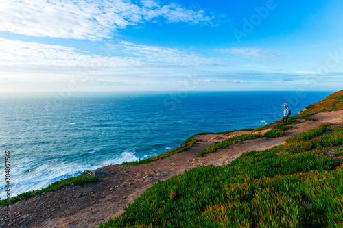 Sintra, Portugal 27 Jan, 2018 - man on high bank with green grass and hiking trails and a view of the blue sea