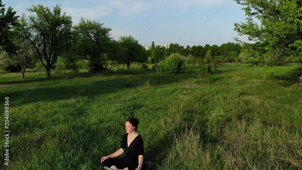 Aerial drone view of a young meditating outdoors on the grass in the park on summer evening during sunset. Aerial drone view of a young meditating outdoors on the grass in the park on summer evening during sunset.