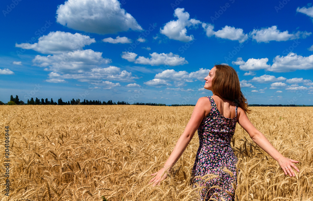 Naklejka premium Beautiful happy young woman in golden wheat field with cloudy blue sky background, copy space. Girl in spikes of ripe wheat field under blue sky