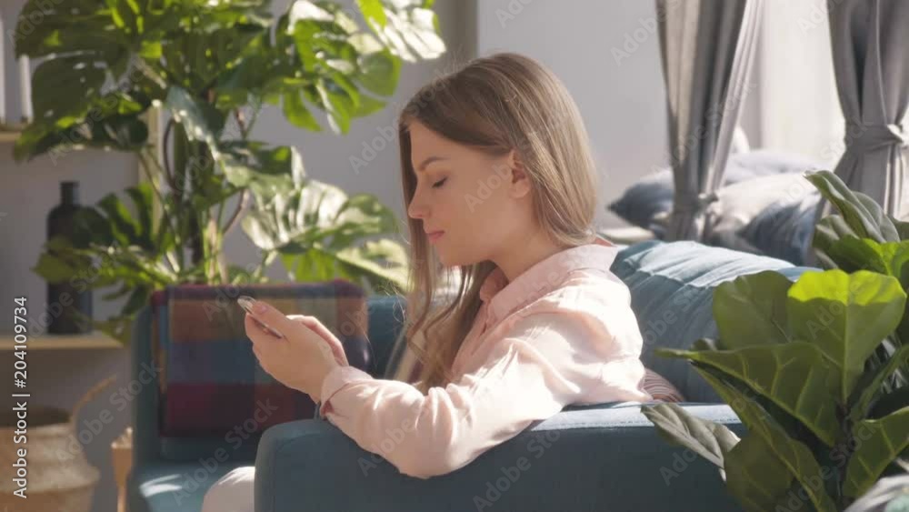 Happy attractive young Caucasian blonde girl texts scrolls and uses app on smartphone cheerfully laughing on a cozy coach in an airy sunny room decorated in elegant Scandinavian style in the morning