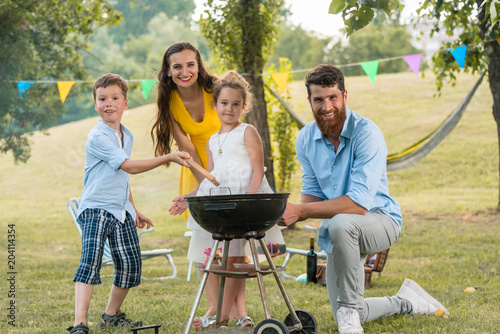 Portrait of a happy family with two children wearing summer casual outfits while posing together outdoors behind a round charcoal barbecue grill