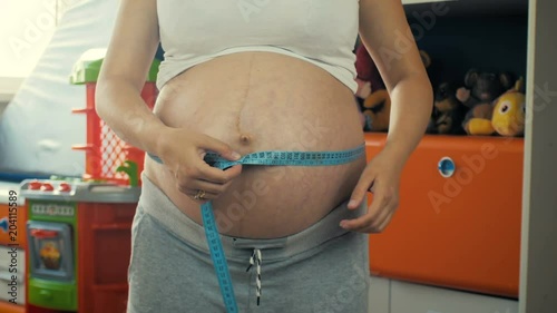 Close-up of a young pregnant girl measures the volume of her belly in the children room, 4K.