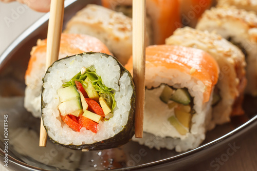 Two chopsticks holding Hosomaki with vegetables and different sushi rolls with seafood on ceramic plate on the background