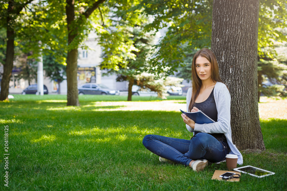 Young brunette asian girl relaxing on a grass and making notes in the park during the day Young brunette asian girl relaxing on a grass and making notes in the park during the day