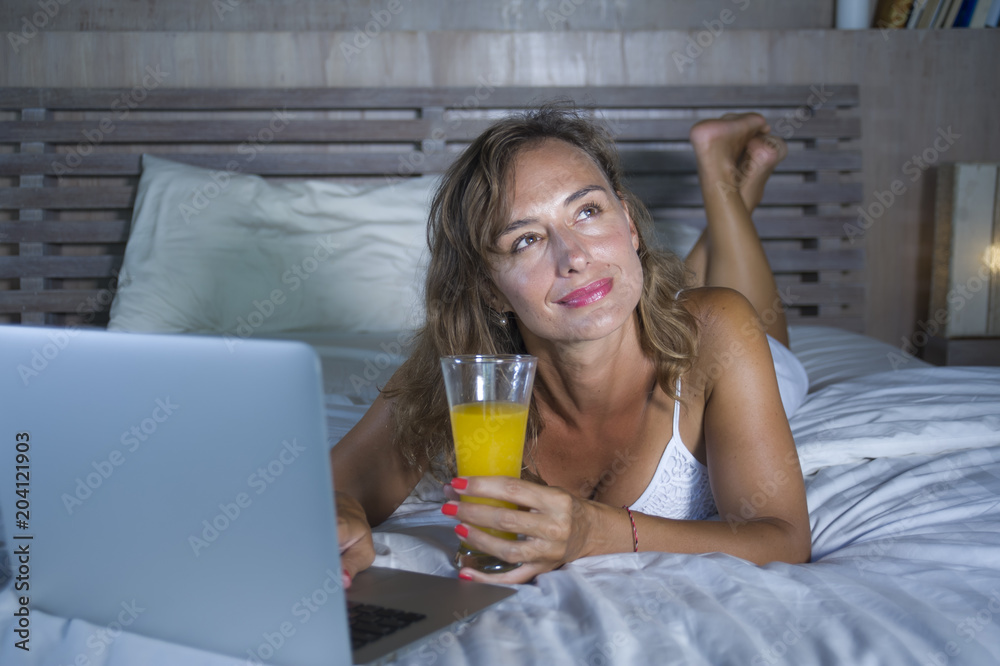 beautiful Caucasian woman 30s lying on bed at night in home bedroom using internet at laptop computer drinking healthy orange juice beautiful Caucasian woman 30s lying on bed at night in home bedroom using internet at laptop computer drinking healthy orange juice